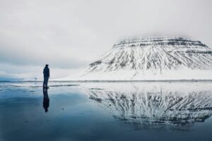 A person standing in front of Kirkjufell mountain in Iceland covered in snow with a reflection in the water